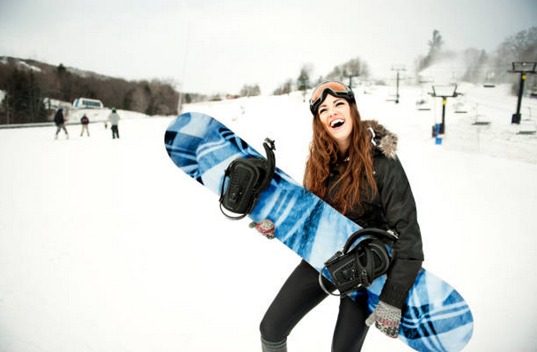 Femme sur une piste de ski qui tient à skateboard en souriant