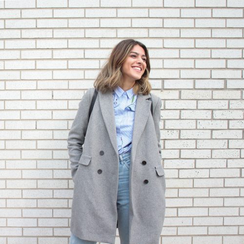 femme portant un jean une chemise bleue et un manteau long gris clair devant un mur en briques blanches